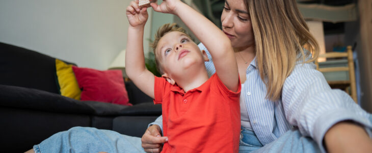 Young boy raising his arms while holding a wooden block with his mother watching him